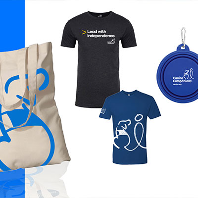 Tote bag with handles bent forward, black t-shirt with white print “lead with independence,” blue shirt with a logo of person and a dog, collapsible water bowl with logo of person and dog